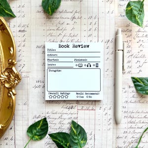 May include: A white square notepad with black printed text titled Book Review, featuring fields for Title, Author, Started, Finished, Genre, Thoughts, and an Overall Rating section with five stars and a Would Recommend section with Yes or No options.