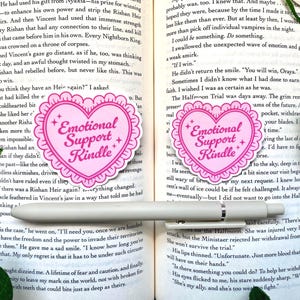 K&ouml;nnte beinhalten: Zwei rosa herzf&ouml;rmige Aufkleber mit dem Text "Emotional Support Kindle". Die Aufkleber haben einen gewellten Rand und Sternakzente. Die Aufkleber befinden sich auf einem Buch mit einem Stift.