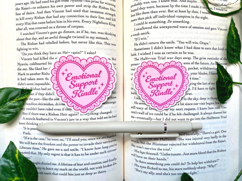 K&ouml;nnte beinhalten: Zwei rosa herzf&ouml;rmige Aufkleber mit dem Text "Emotional Support Kindle". Die Aufkleber haben einen gewellten Rand und Sternakzente. Die Aufkleber befinden sich auf einem Buch mit einem Stift.
