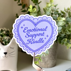 K&ouml;nnte beinhalten: Ein herzf&ouml;rmiger Aufkleber in Hellviolett mit dem Text "Emotional Support Kindle". Der Aufkleber hat einen gewellten Rand und Sterne. Er wird von einer Pinzette gehalten. Dekoratives Objekt.