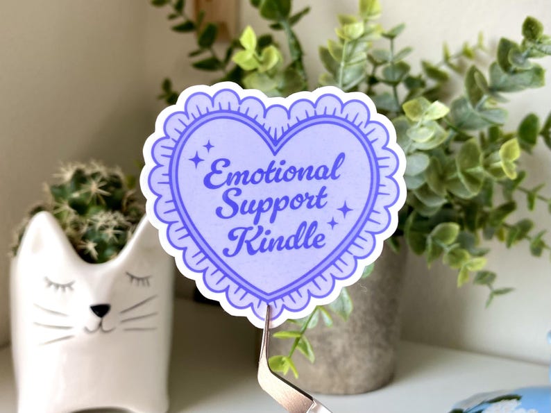 K&ouml;nnte beinhalten: Ein herzf&ouml;rmiger Aufkleber in Hellviolett mit dem Text "Emotional Support Kindle". Der Aufkleber hat einen gewellten Rand und Sterne. Er wird von einer Pinzette gehalten. Dekoratives Objekt.