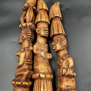 Pode incluir: Três esculturas verticais em madeira com figuras humanas empilhadas num arranjo tipo totem. Cada escultura exibe traços faciais detalhados, vestuário texturizado e toucados pontiagudos com um acabamento natural de grão de madeira castanho.