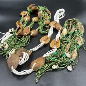 May include: Two multi-strand necklaces featuring green seed beads, white seed beads, and brown natural seed pods. The necklaces are accented with small white cowrie shells and green cylindrical beads, arranged in a layered, cascading design.