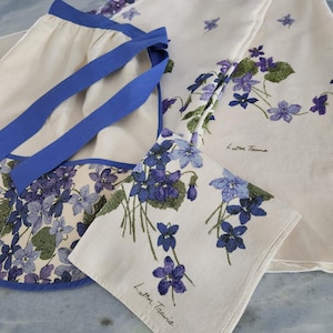 Könnte beinhalten: Ein Set aus cremefarbenen Leinen und einer Schürze, alle mit einem Blumendesign aus lila und blauen Veilchen mit grünen Blättern. Die Schürze hat blaue Paspeln und Bindebänder. Der Text "Lutter Travis" ist auf die Leinen gedruckt.