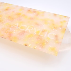 May include: A close-up of a sheet of translucent acrylic with a speckled pattern of yellow and pink. The word "ACRYLIC" is printed in white on the sheet.