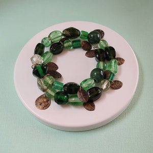 May include: A beaded bracelet featuring a variety of green beads in different shapes and shades, accented with bronze-colored leaf charms and a decorative pendant. The bracelet is displayed on a white circular surface.