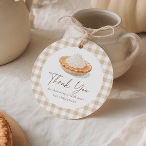 May include: A round gift tag with a tan and white gingham pattern. The tag features a watercolor illustration of a pie slice topped with whipped cream. The text reads "Thank You" and "for showering us with love! THE ANDERSONS."