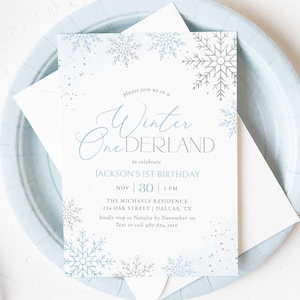 May include: A light blue invitation card with the text "Winter OneDERLAND" and snowflake designs. The invitation is for Jackson's 1st birthday, with the date November 30th at 1 PM. The card is on a light blue plate with a white envelope.