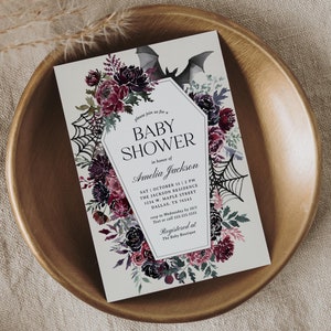 May include: A white invitation card with a black and white illustration of a bat and a spider web. The card has a floral design with red, purple, and green flowers. The text on the card reads "Please join us for a Baby Shower in honor of Amelia Jackson. Sat October 15 | 2 PM The Jackson Residence 1234 W. Maple Street Dallas, TX 76543 RSVP to Wednesday by 30/1 Text or call 555.555.5555 Registered at The Baby Boutique."