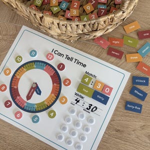 Pode incluir: Um quadro educativo I Can Tell Time com mostrador de relógio, ponteiros móveis e pontos de velcro para combinar peças numéricas. O conjunto inclui peças retangulares coloridas com números e palavras escritas, guardadas num cesto de vime.