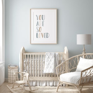 Puede incluir: Una cuna blanca con una colcha blanca y gris, un caballito de balanc&iacute;n y una silla de mimbre blanca con un coj&iacute;n blanco. Un cuadro enmarcado con el texto "YOU ARE SO LOVED" cuelga de la pared sobre la cuna.
