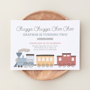 Op de afbeelding: Een verjaardagsuitnodiging met de tekst "Chugga Chugga Choo Choo" en "Grayson wordt twee!" Een cartoon trein illustratie in blauw, geel en rood is te zien onder de tekst. De uitnodiging ligt op een houten bord.