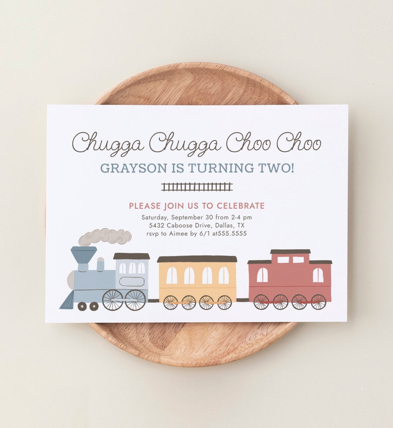May include: A birthday invitation with the text "Chugga Chugga Choo Choo" and "Grayson is turning two!" A cartoon train illustration in blue, yellow, and red is featured below the text. The invitation is on a wooden plate.