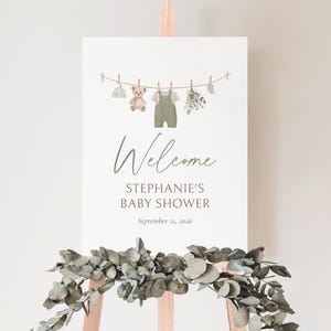 K&ouml;nnte beinhalten: Ein Willkommensschild f&uuml;r eine Babyparty auf einer Holzstaffelei. Das Schild zeigt eine Aquarellillustration von Babykleidung, einem Teddyb&auml;r und Gr&uuml;nzeug, das an einer W&auml;scheleine h&auml;ngt. Auf dem Schild steht "Welcome, Stephanie's Baby Shower, September 21, 2026."