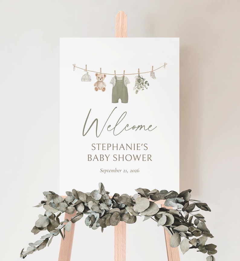 K&ouml;nnte beinhalten: Ein Willkommensschild f&uuml;r eine Babyparty auf einer Holzstaffelei. Das Schild zeigt eine Aquarellillustration von Babykleidung, einem Teddyb&auml;r und Gr&uuml;nzeug, das an einer W&auml;scheleine h&auml;ngt. Auf dem Schild steht "Welcome, Stephanie's Baby Shower, September 21, 2026."