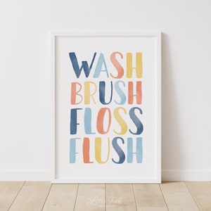 May include: A white framed print with the words "WASH BRUSH FLOSS FLUSH" in colorful watercolor lettering. The words are stacked vertically and are in shades of blue, yellow, orange, and pink.