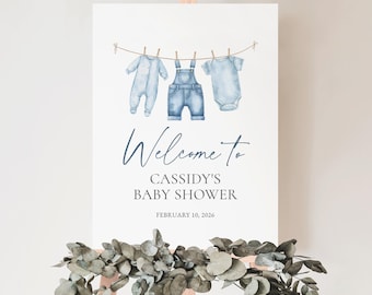 May include: A baby shower sign on an easel with the text "Welcome to Cassidy's Baby Shower" and the date February 10, 2026. The sign features watercolor illustrations of baby clothes hanging on a clothesline. Eucalyptus greenery is at the base.