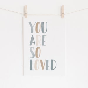 Puede incluir: Una tarjeta blanca con las palabras "YOU ARE SO LOVED" escritas en letras de acuarela azul y marr&oacute;n. La tarjeta est&aacute; colgada de una cuerda con pinzas para la ropa.
