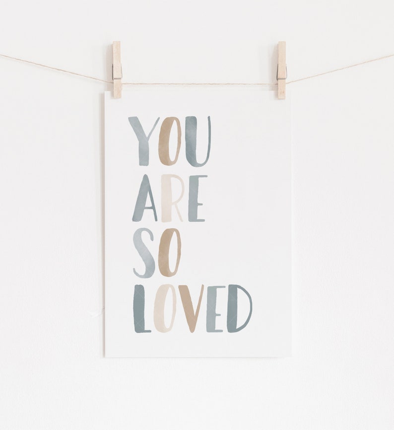 Puede incluir: Una tarjeta blanca con las palabras "YOU ARE SO LOVED" escritas en letras de acuarela azul y marr&oacute;n. La tarjeta est&aacute; colgada de una cuerda con pinzas para la ropa.