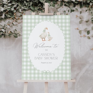 Könnte beinhalten: Ein Babyparty-Schild auf einer Holzstaffelei. Das Schild hat einen hellgrünen Vichy-Muster-Hintergrund mit einem ovalen Rahmen. Im Inneren des Rahmens befinden sich zwei Gänse mit Schleifen und der Text "Welcome to Cassidy's Baby Shower August 25, 2027."