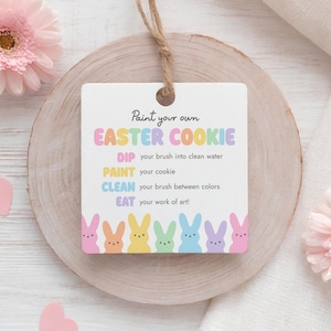 May include: A square, white sign with the text "Paint your own EASTER COOKIE" in pastel colours. Instructions are below the title. A row of colourful bunny illustrations is at the bottom. The sign hangs from a natural twine string.