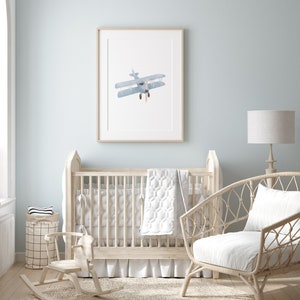 May include: A white crib with a white and gray quilt, a rocking horse, and a wicker chair with a white cushion. A framed print of a blue airplane hangs on the wall above the crib.