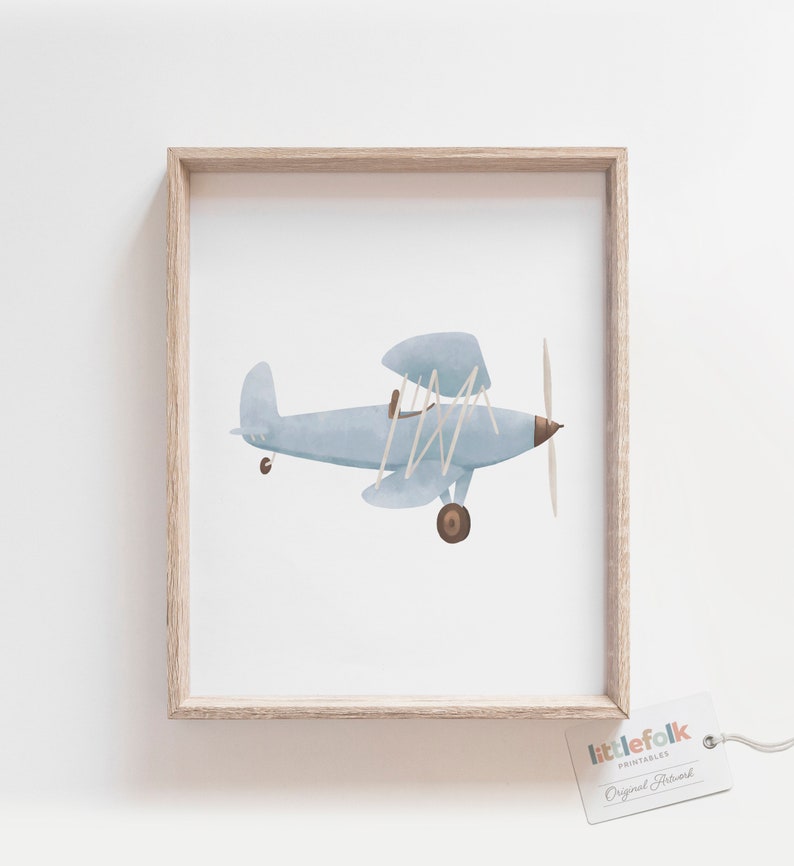 K&ouml;nnte beinhalten: Eine Aquarell-Illustration eines hellblauen Flugzeugs mit braunem Propeller und R&auml;dern. Das Flugzeug ist in einem hellen Holzrahmen gerahmt.