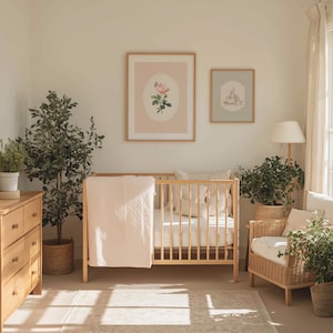 May include: A nursery scene with a light wood crib, a dresser, and a wicker chair. Two framed prints hang on the wall, one with a rose illustration. A pink blanket drapes over the crib. Plants and a lamp complete the cozy, neutral-toned room.
