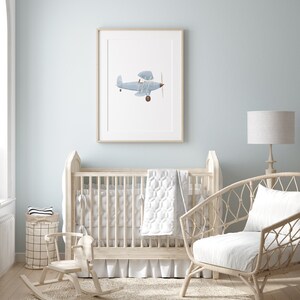 K&ouml;nnte beinhalten: Ein hellblaues Holzbabybett mit einem wei&szlig;en Bettrock und einer wei&szlig;en gesteppten Decke. Ein Schaukelpferd und ein Korbstuhl mit einem wei&szlig;en Kissen stehen im Raum. Ein gerahmtes Bild eines blauen Flugzeugs h&auml;ngt an der Wand.