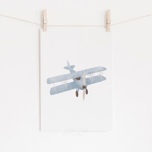 May include: A printable wall art design featuring a blue watercolor illustration of a vintage airplane with a brown propeller.
