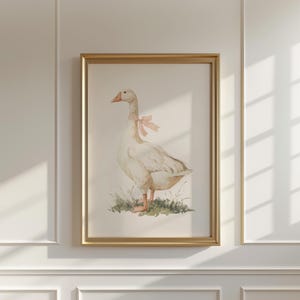 May include: A framed watercolor print of a goose with a pink bow around its neck. The goose stands on a patch of green grass. The artwork is in a gold frame and hangs on a white wall.