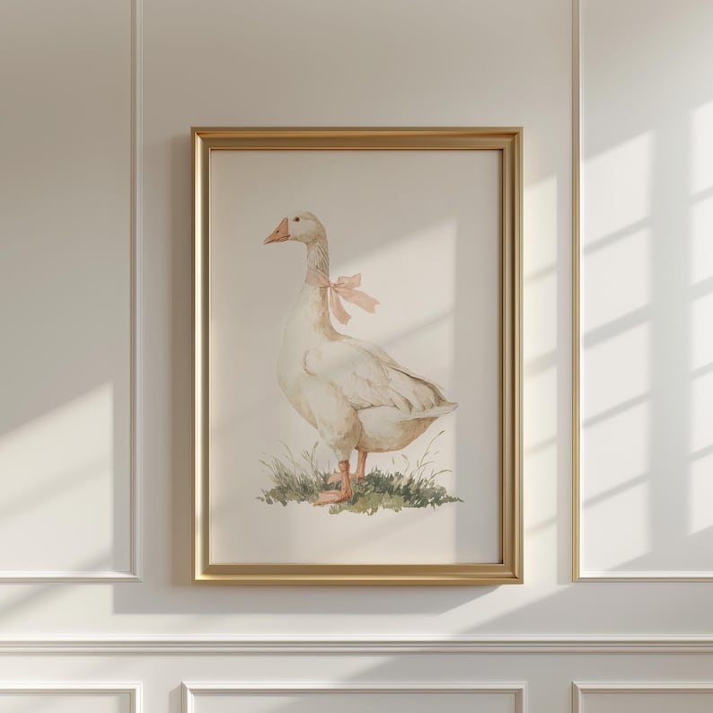 May include: A framed watercolor print of a goose with a pink bow around its neck. The goose stands on a patch of green grass. The artwork is in a gold frame and hangs on a white wall.