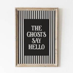 Könnte beinhalten: Gerahmtes Wandbild mit schwarz-weiß gestreiftem Hintergrund und schwarzer Mitte. Der Text "THE GHOSTS SAY HELLO" ist in weißer, stilisierter Schrift dargestellt. Der Rahmen hat eine helle, natürliche Holzfarbe.