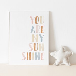 May include: A white framed print with the text "You are my sunshine" in colorful watercolor letters. The letters are in shades of pink, orange, blue, and yellow.