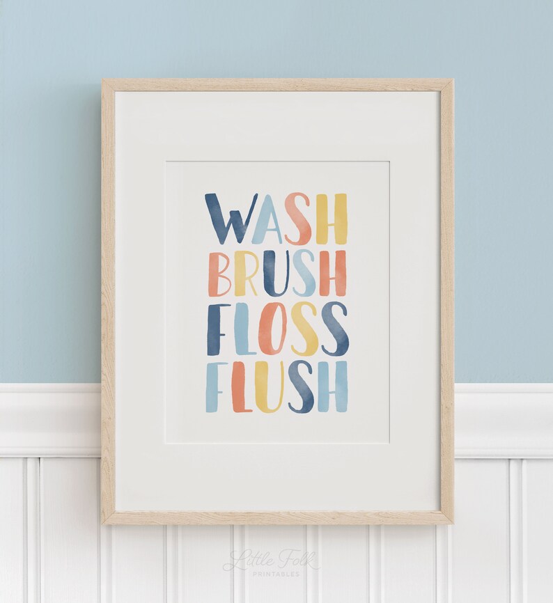 May include: A framed print with the words "WASH BRUSH FLOSS FLUSH" in colorful, watercolor-style lettering. The print is on a white background and has a light wood frame.