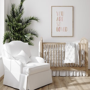 K&ouml;nnte beinhalten: Ein wei&szlig;er Kinderzimmer-Schaukelstuhl mit einem wei&szlig;en Kissen steht neben einem Holz-Kinderbett mit einer wei&szlig;en Steppdecke. Ein gerahmtes Bild mit dem Text "You are so loved" h&auml;ngt an der Wand hinter dem Kinderbett.