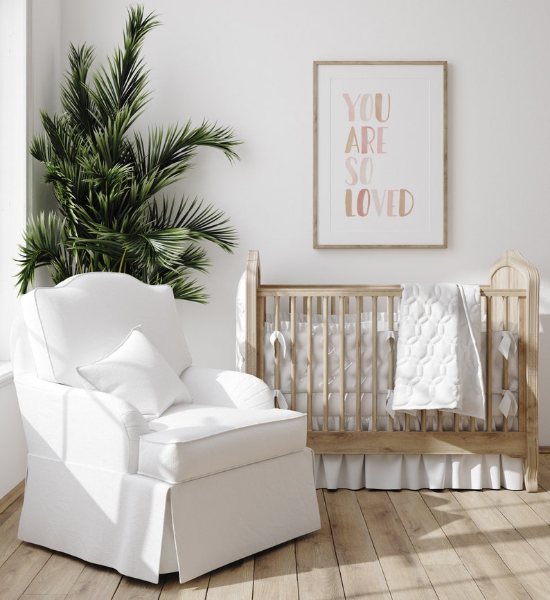 K&ouml;nnte beinhalten: Ein wei&szlig;er Kinderzimmer-Schaukelstuhl mit einem wei&szlig;en Kissen steht neben einem Holz-Kinderbett mit einer wei&szlig;en Steppdecke. Ein gerahmtes Bild mit dem Text "You are so loved" h&auml;ngt an der Wand hinter dem Kinderbett.