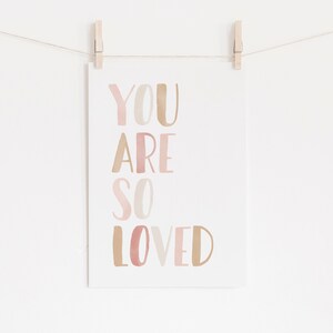 K&ouml;nnte beinhalten: Eine wei&szlig;e Karte mit dem Text "YOU ARE SO LOVED" in rosa und braunen Aquarellbuchstaben. Die Karte h&auml;ngt an einer Schnur mit W&auml;scheklammern.