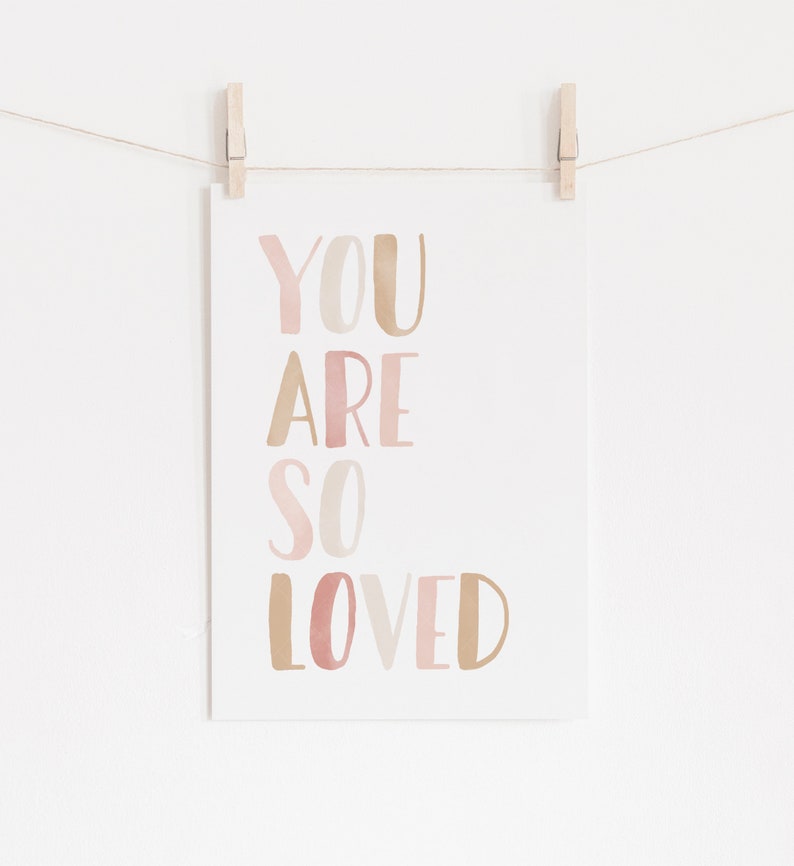 K&ouml;nnte beinhalten: Eine wei&szlig;e Karte mit dem Text "YOU ARE SO LOVED" in rosa und braunen Aquarellbuchstaben. Die Karte h&auml;ngt an einer Schnur mit W&auml;scheklammern.