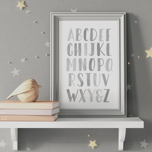 May include: A framed print with a gray background and white letters spelling out the alphabet in a watercolor style. The frame is silver and the print is on a white shelf with a gold bird figurine and three books.