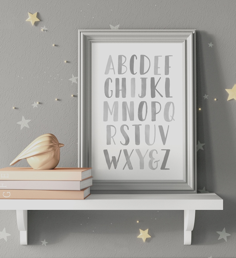 May include: A framed print with a gray background and white letters spelling out the alphabet in a watercolor style. The frame is silver and the print is on a white shelf with a gold bird figurine and three books.