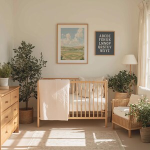 Op de afbeelding: Een lichte kinderkamer met een houten wieg, commode en fauteuil. Een ingelijste honkbalveldprint en een alfabetkaart hangen aan de muur. Een roze deken hangt over de wieg. Planten in manden en potten voegen groen toe.