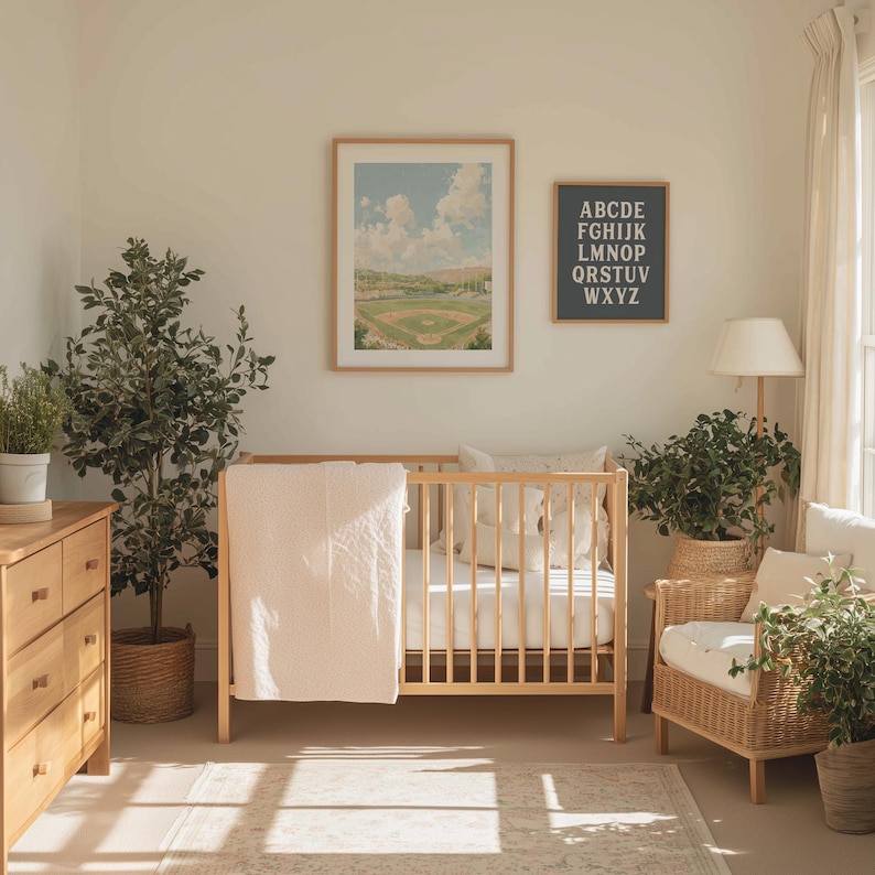 Op de afbeelding: Een lichte kinderkamer met een houten wieg, commode en fauteuil. Een ingelijste honkbalveldprint en een alfabetkaart hangen aan de muur. Een roze deken hangt over de wieg. Planten in manden en potten voegen groen toe.