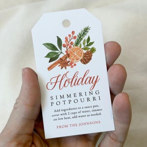 May include: A white rectangular gift tag with a hole at the top, featuring a watercolor illustration of orange slices, cinnamon sticks, pine needles, and berries. Text reads: Holiday SIMMERING POTPOURRI. Add ingredients to a sauce pan, cover with 2 cups of water, simmer on low heat, add water as needed. FROM THE JOHNSONS.