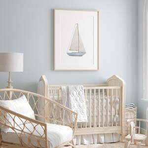 May include: A white cot with a white and grey quilt and a white frilled skirt. A wicker chair with a white cushion sits in front of the cot. A framed watercolour painting of a sailboat hangs on the wall behind the cot.