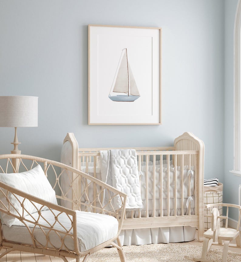 May include: A white cot with a white and grey quilt and a white frilled skirt. A wicker chair with a white cushion sits in front of the cot. A framed watercolour painting of a sailboat hangs on the wall behind the cot.
