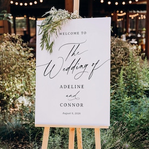 May include: A white canvas wedding sign on a wooden easel. The sign reads "WELCOME TO The Wedding of ADELINE and CONNOR August 9, 2026" in elegant black script. Greenery and white flowers adorn the top of the sign, with a blurred outdoor background.