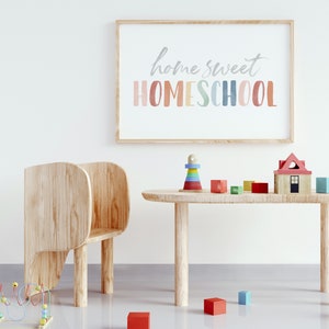 May include: A light wood table with a wooden chair and a variety of colorful wooden toys. The toys include a house, a stacking tower, and blocks. The wall behind the table has a framed print that says "home sweet homeschool" in colorful letters.