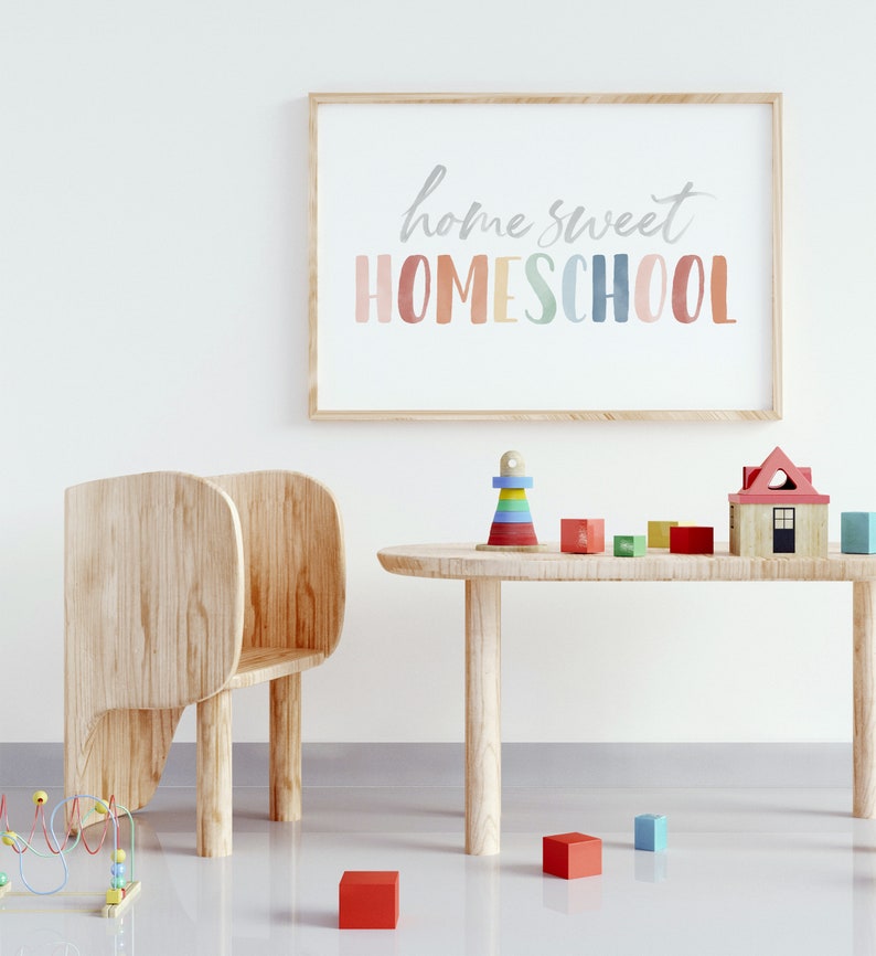 May include: A light wood table with a wooden chair and a variety of colorful wooden toys. The toys include a house, a stacking tower, and blocks. The wall behind the table has a framed print that says "home sweet homeschool" in colorful letters.