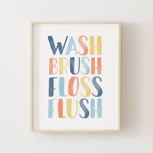 May include: A framed print with the words "WASH BRUSH FLOSS FLUSH" in colorful, watercolor-style lettering. The words are stacked vertically and are in shades of blue, yellow, and orange. The print is framed in a light wood frame.